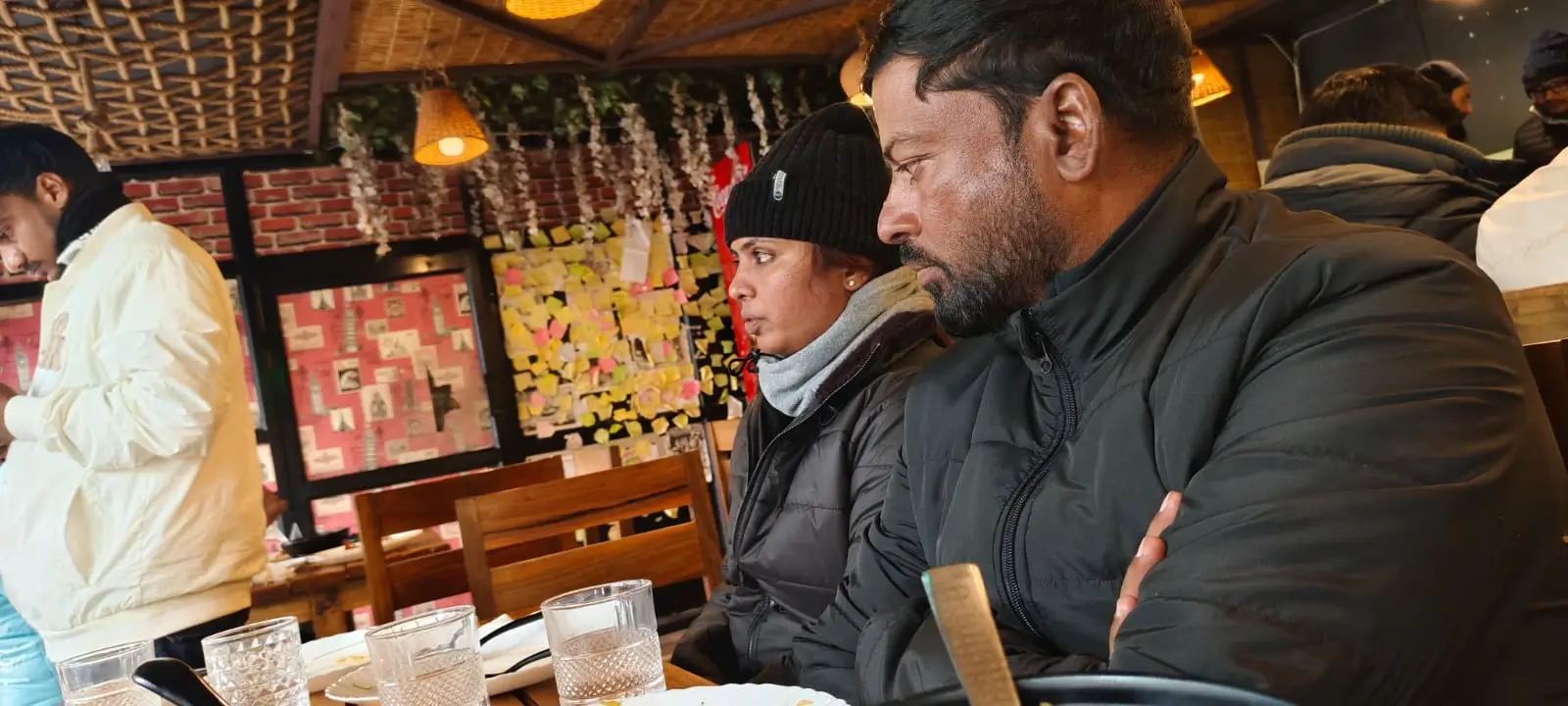 Two individuals seated at a table in a restaurant, wearing winter clothing and looking thoughtfully to the side.