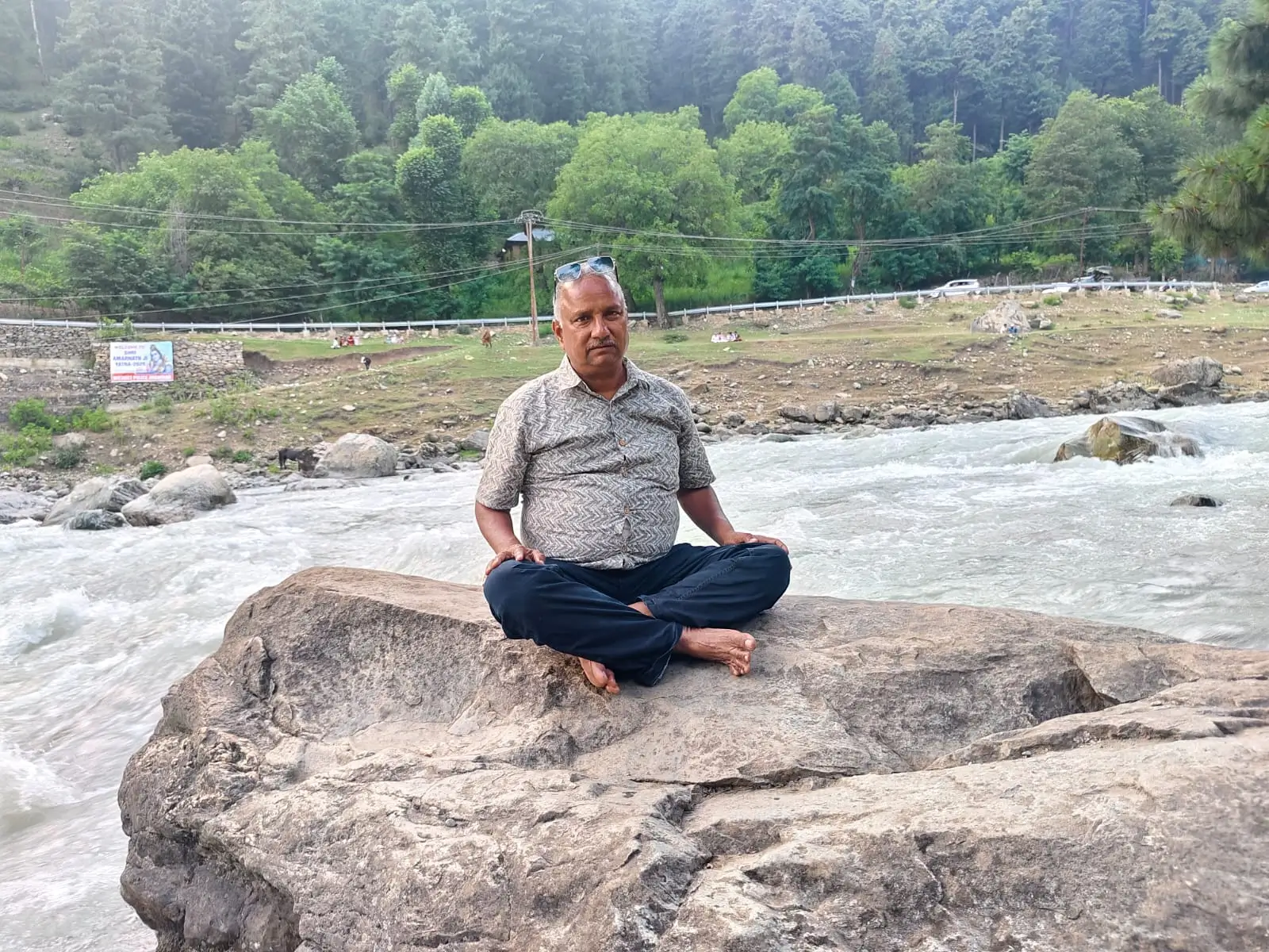 Man sitting cross-legged on a rock by a flowing river in a natural setting.