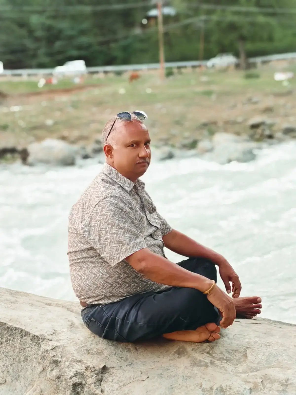 Man sitting on a rock by a river, wearing a patterned shirt and sunglasses, with water flowing in the background.