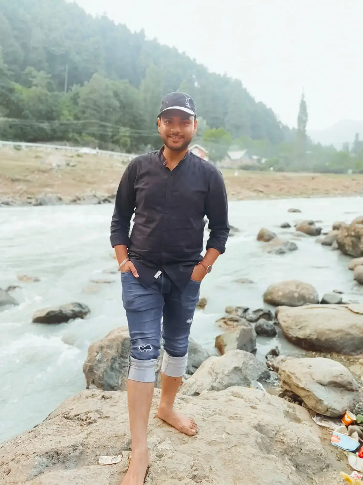 Man standing on rocks by a river in Kashmir, wearing a black shirt and denim jeans, with a cap and a serene landscape in the background.