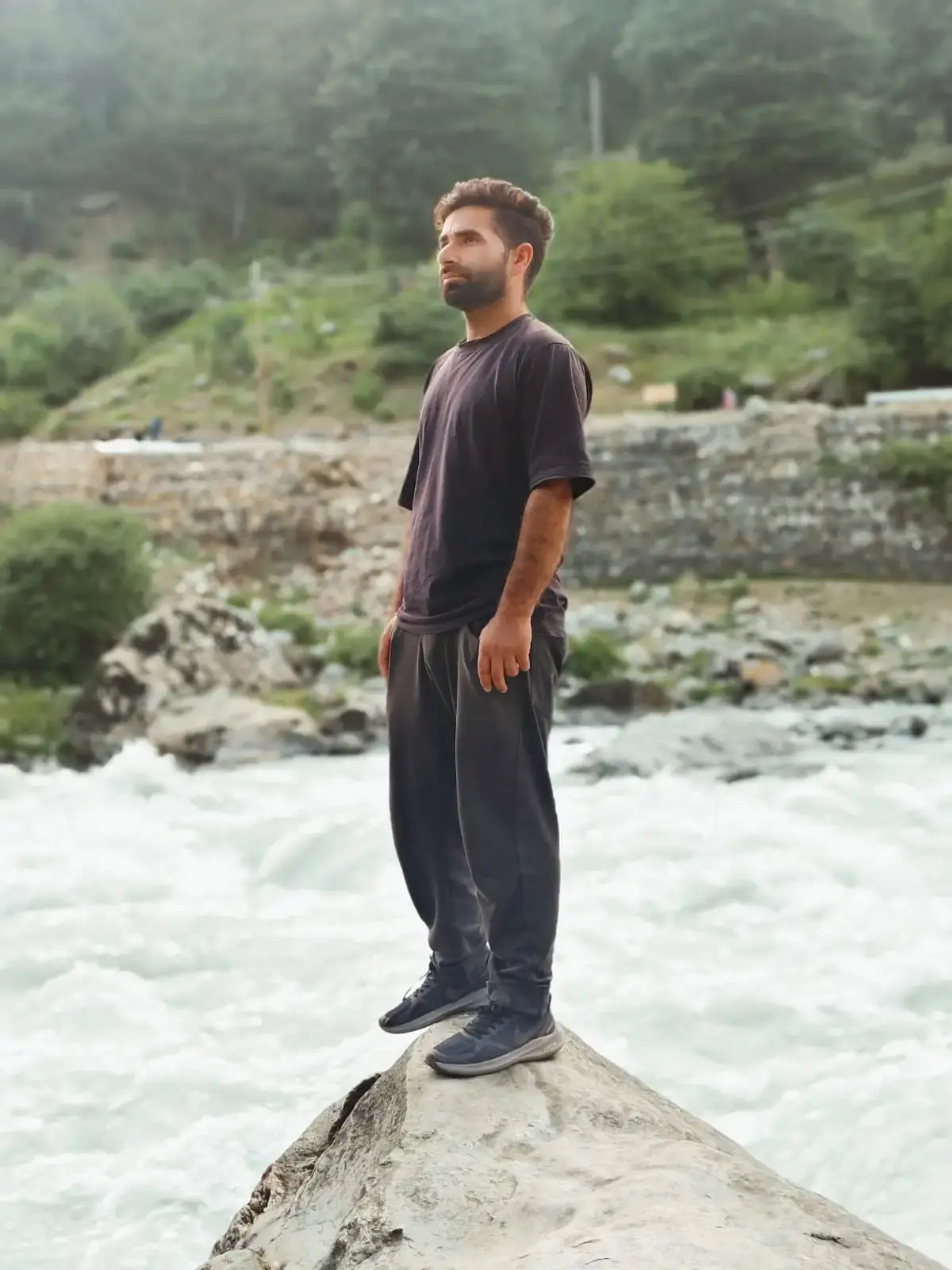 Man standing on a rock by a river, wearing a black t-shirt and dark pants, with flowing water in the background.