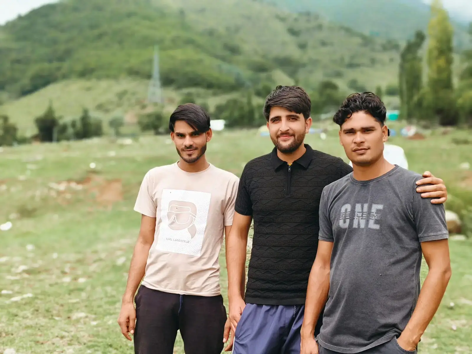 Three young men standing together outdoors in a green landscape with hills in the background.